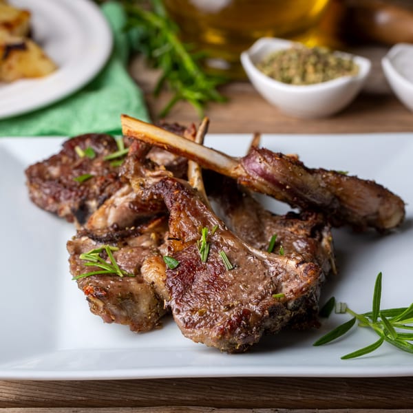 Grilled lamb chops with rosemary on a white plate, with spices and potatoes in the background.
