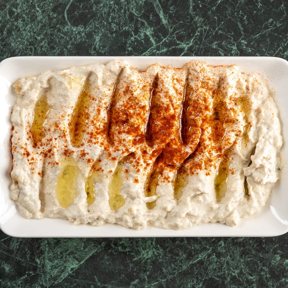 A white rectangular plate of creamy baba ghanoush, drizzled with olive oil and sprinkled with paprika.