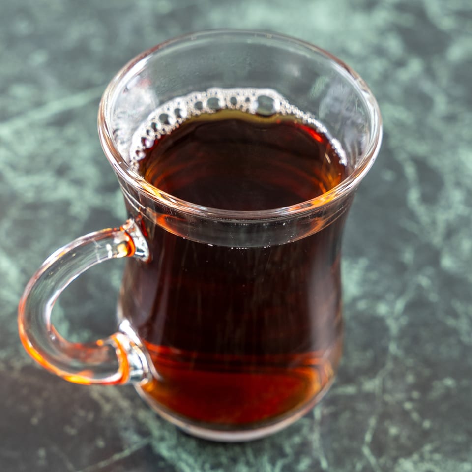 A glass of Turkish tea with a handle, filled with dark amber liquid and a few bubbles.