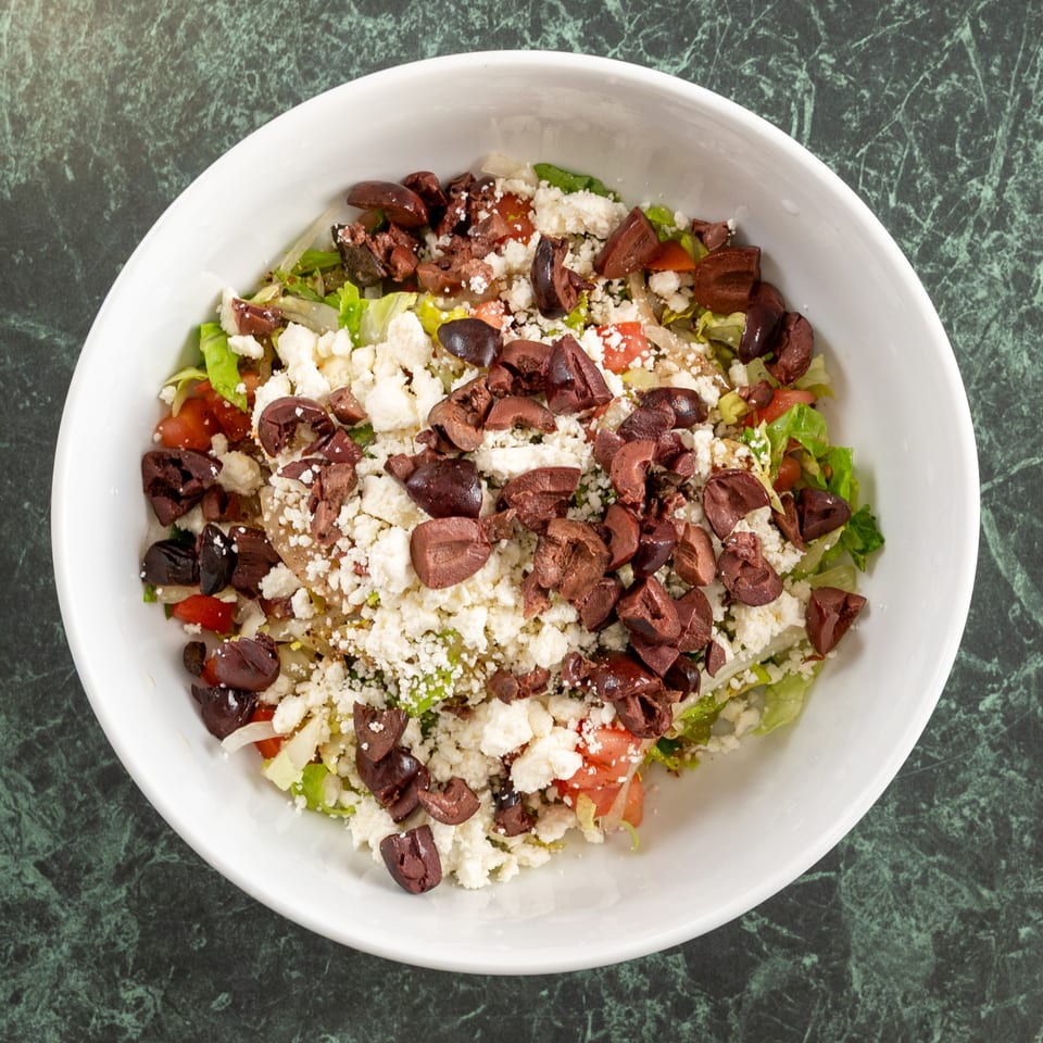 A white bowl filled with a Mediterranean salad, topped with feta cheese and black olives.