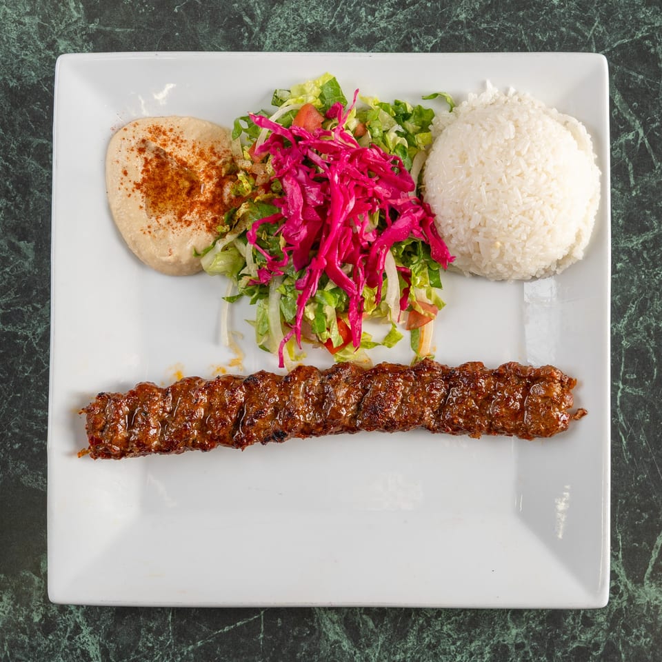 Adana kebab with rice, hummus, and salad with red cabbage, served on a white plate.